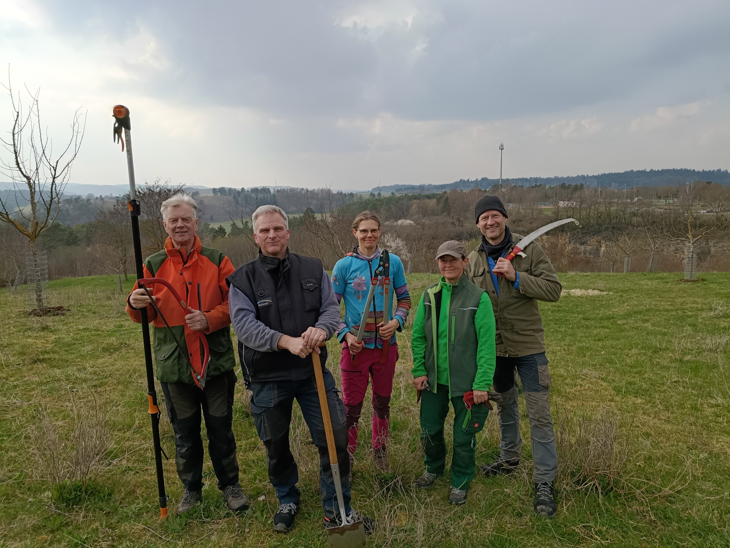 Eine Gruppe mit Personen hat Obstbaumwerkzeuge in der Hand und schaut auf einer Streuobstwiese in Richtung Kamera